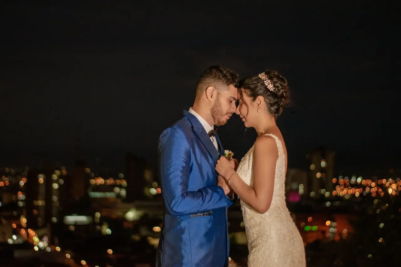 Noivos de mãos dadas e com as testas encostadas durante ensaio fotográfico noturno, com luzes da cidade desfocadas ao fundo.