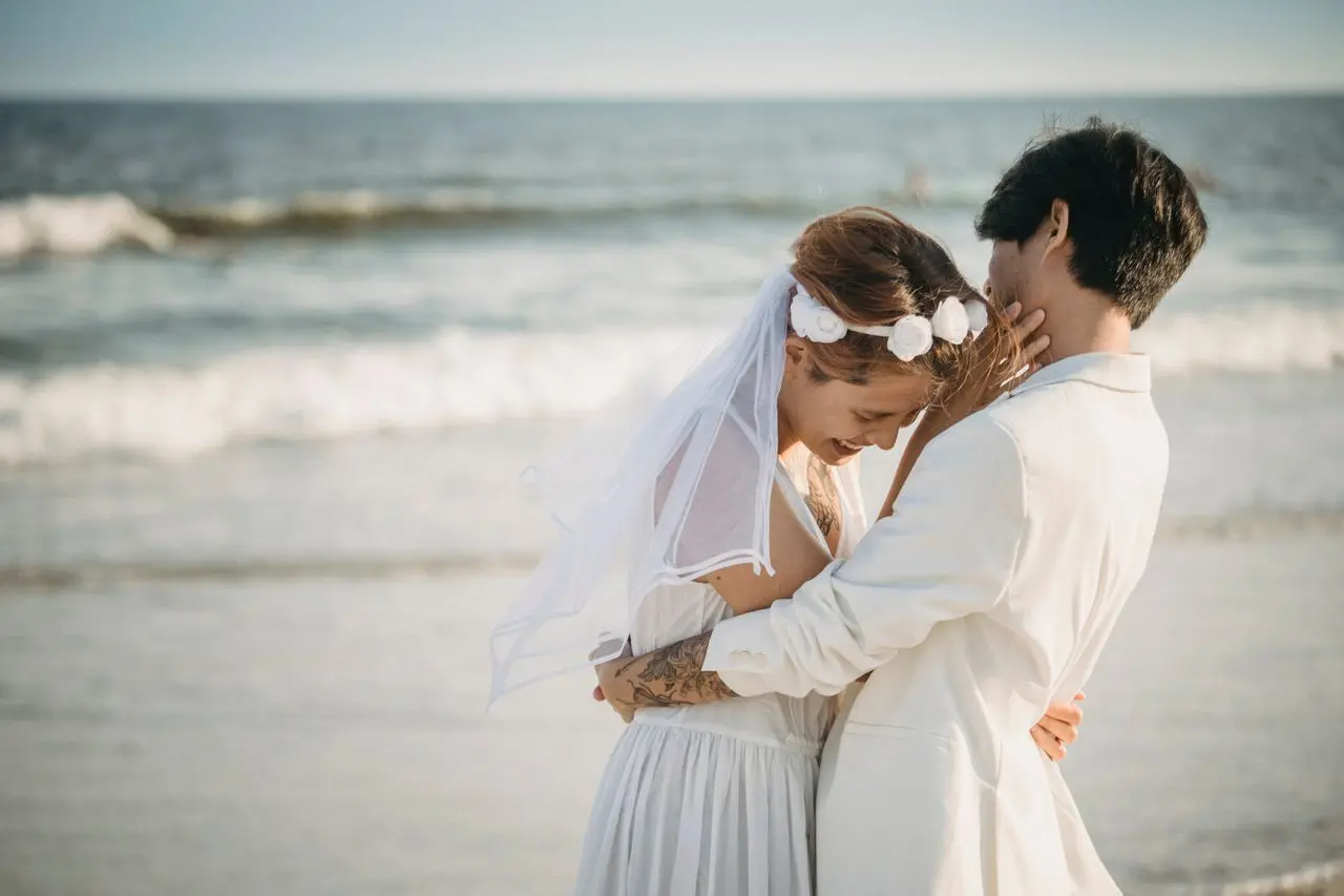 Casal de noivos se abraçando durante casamento na praia, com o mar ao fundo.