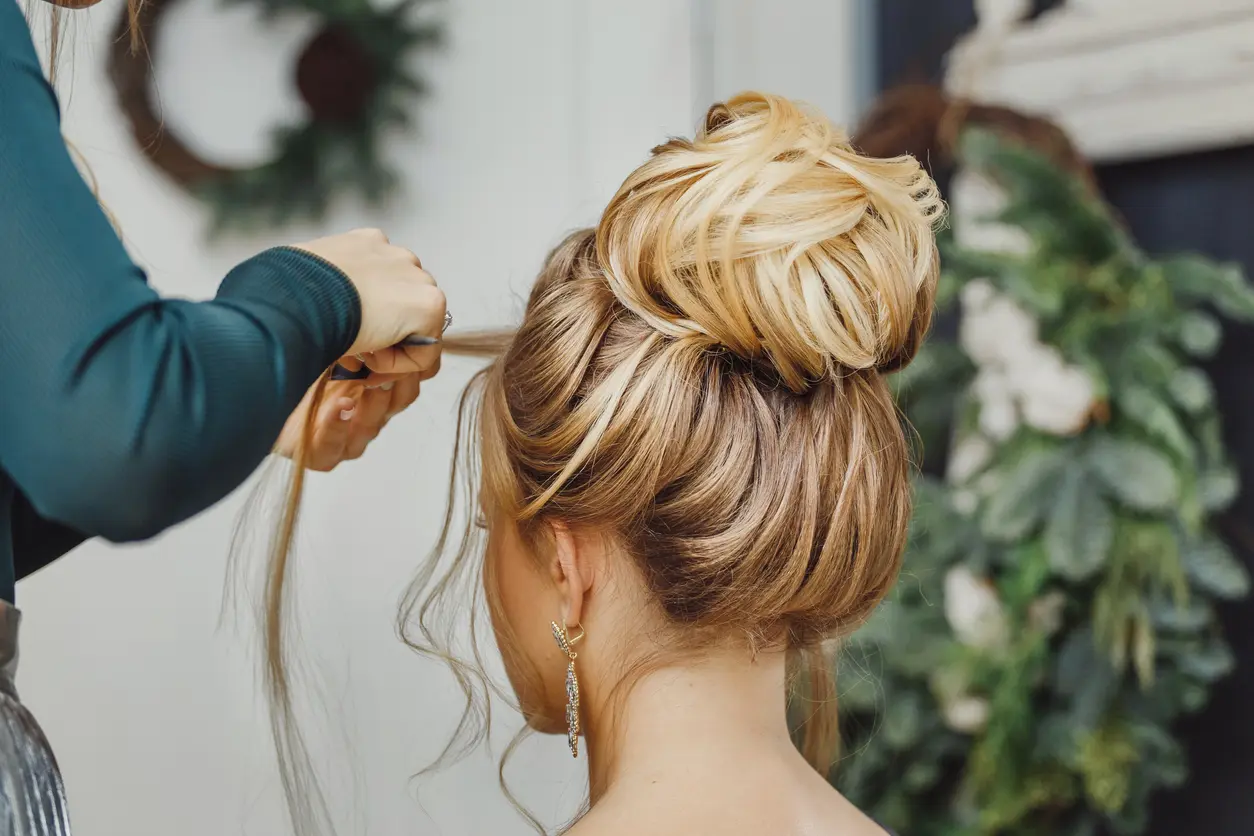 Penteado de noiva simples: solto, preso ou semi-preso para cabelo longo e curto Imagem mostra uma mulher em um salão de beleza, fazendo um penteado com a ajuda de um profissional, que trabalha cuidadosamente em seu cabelo
