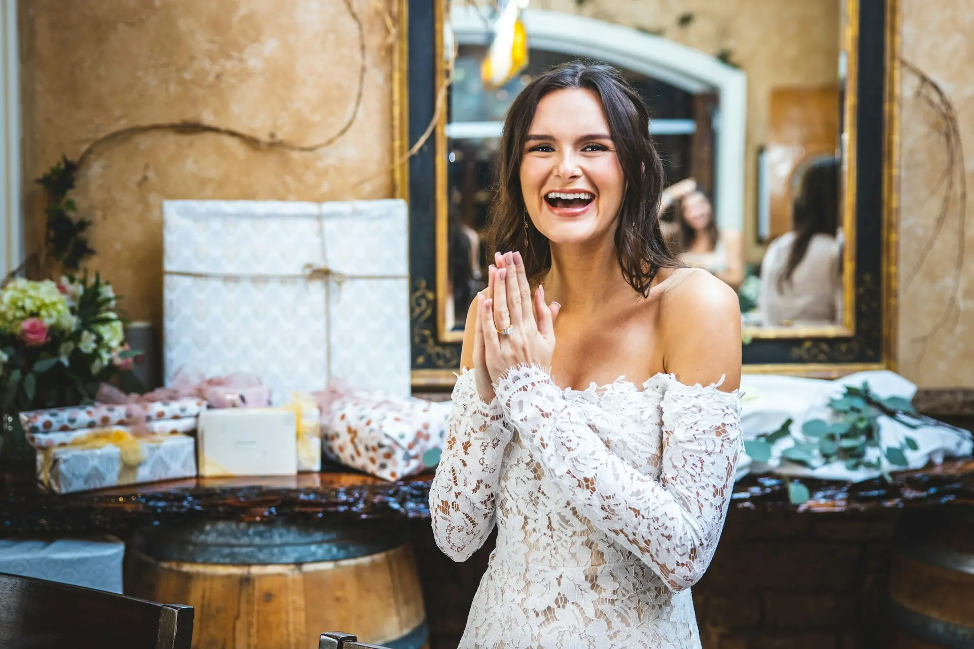 Noiva sorridente com vestido branco de renda e mangas longas, exibindo a aliança durante a abertura dos presentes do chá de panela.