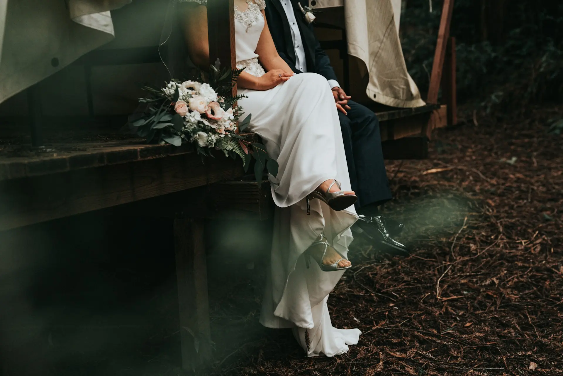 Casal de noivos sentado em uma estrutura de madeira ao ar livre, de mãos dadas, com buquê de flores em tons claros e decoração rústica.