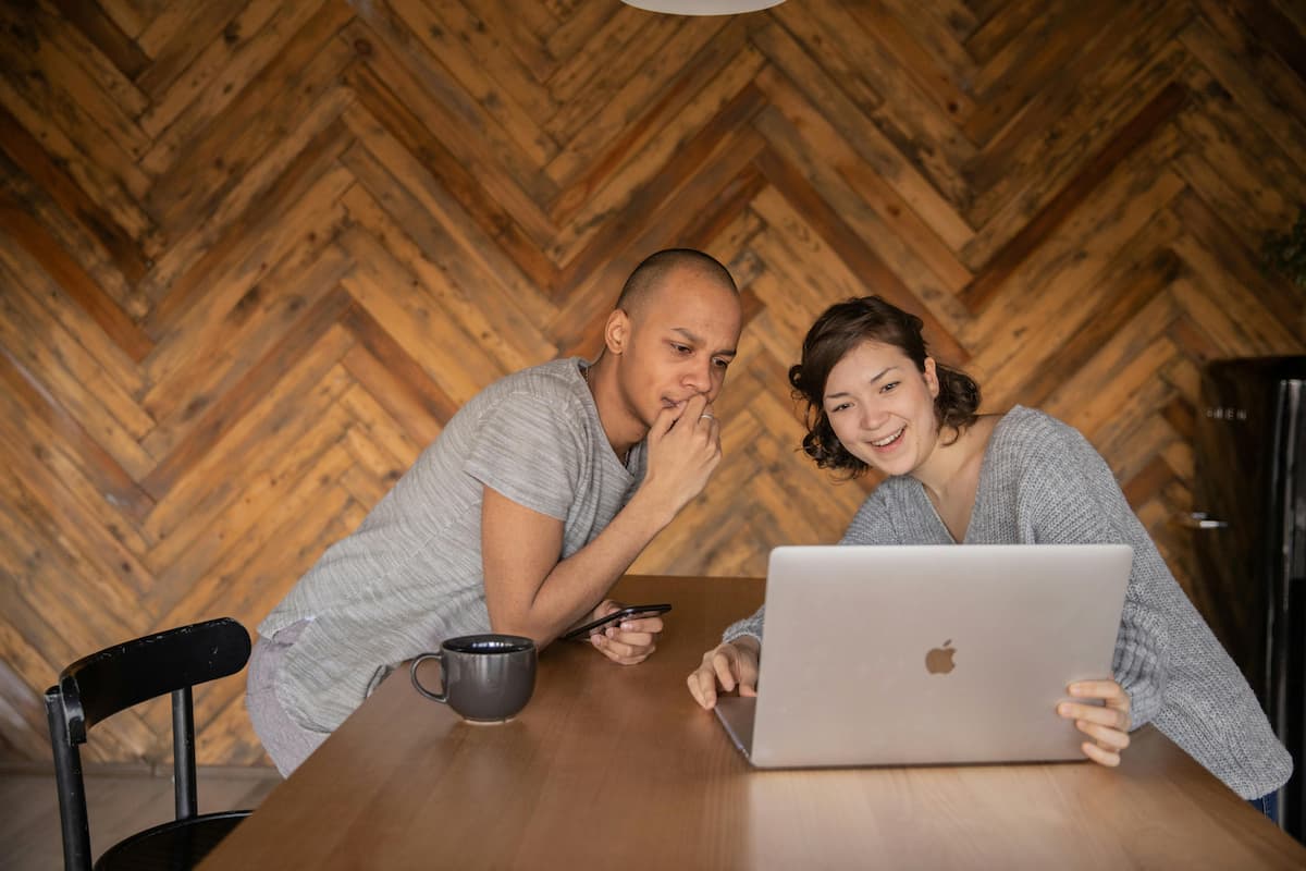 Casal sentado à mesa usando notebook e celular, sorrindo enquanto monta uma lista de casamento divertida em ambiente aconchegante.