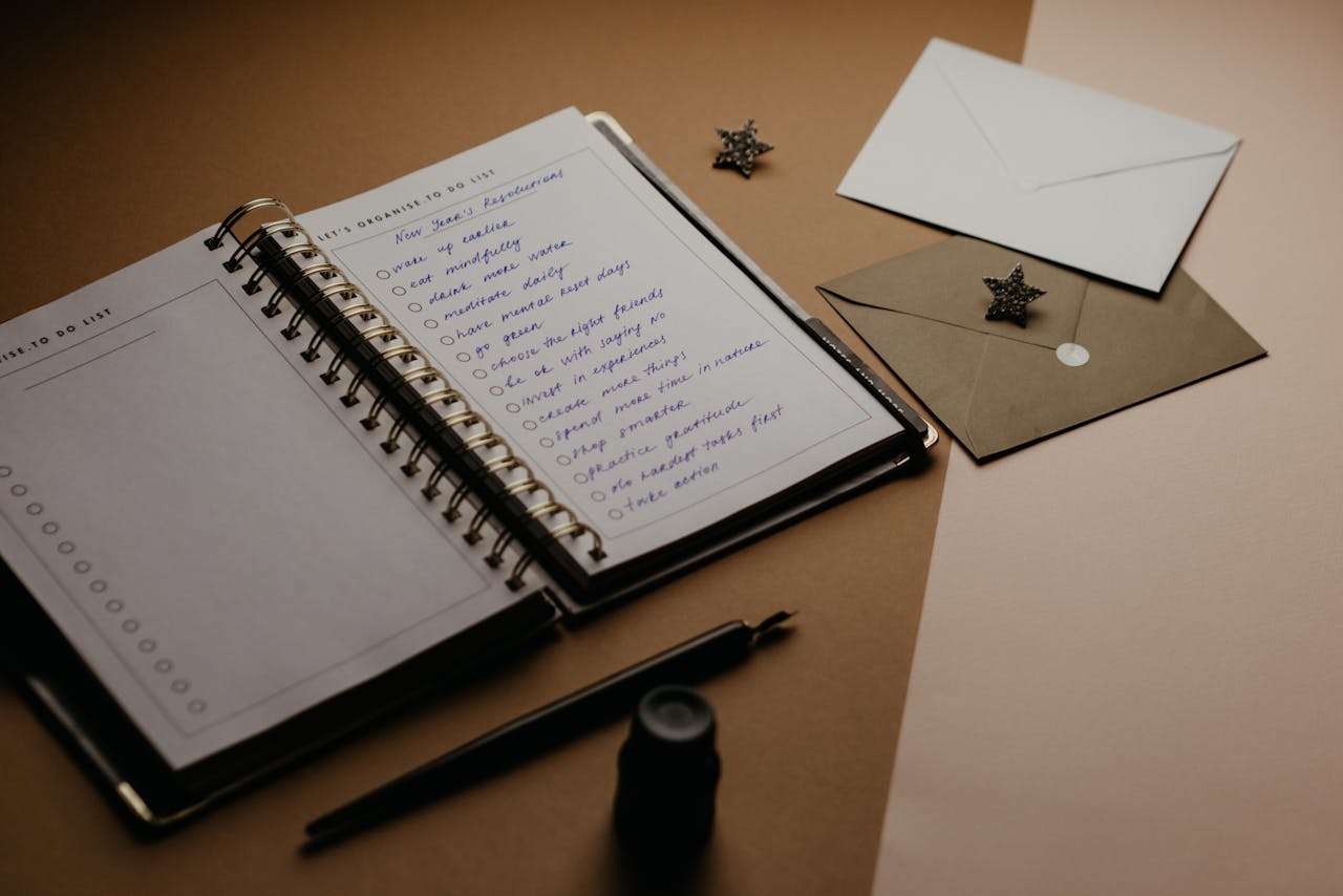 Imagem de um caderno aberto com uma lista para chá de cozinha, ao lado há envelopes de convite e canetas sobre a mesa.