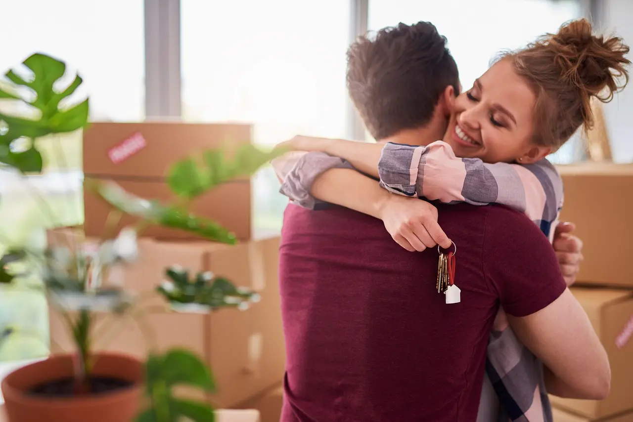 Imagem de um casal se abraçando, com caixas de mudança ao fundo, aparentando ser um casal feliz com a casa nova.
