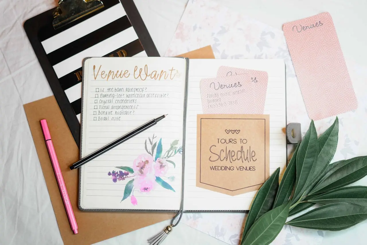 Imagem de um caderno aberto com checklist para casamento, com caneta em cima, cartões escritos, folhas verdes decorativas ao lado e itens de papelaria sobre a mesa.
