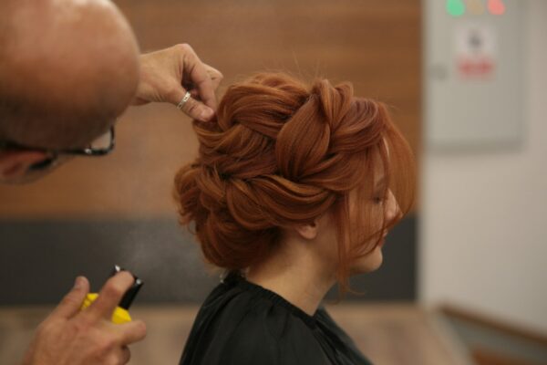 Imagem mostra uma mulher em um salão de beleza, fazendo um penteado com a ajuda de um profissional, que trabalha cuidadosamente em seu cabelo.