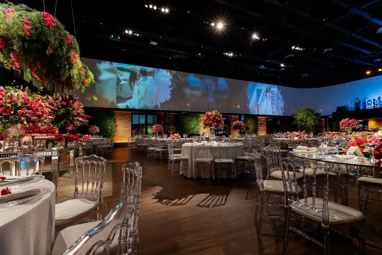 Como criar uma recepção elegante para convidados sem exagerar A imagem mostra um salão de casamento elegante, com mesas decoradas, flores em tons de vermelho e um grande painel ao fundo com fotos do casal.