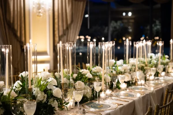 Imagem de um casamento com decoração moderna. Mesa com muitas velas altas, caminho de flores e local iluminado.