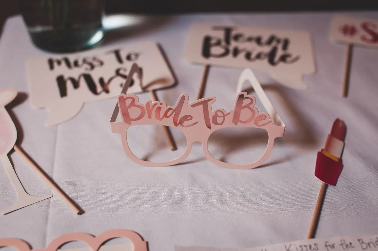 7 ideias para arrecadar dinheiro para o casamento Óculos decorativos de despedida de solteira com a frase “Bride to Be”, cercados por plaquinhas temáticas e um acessório em formato de batom.
