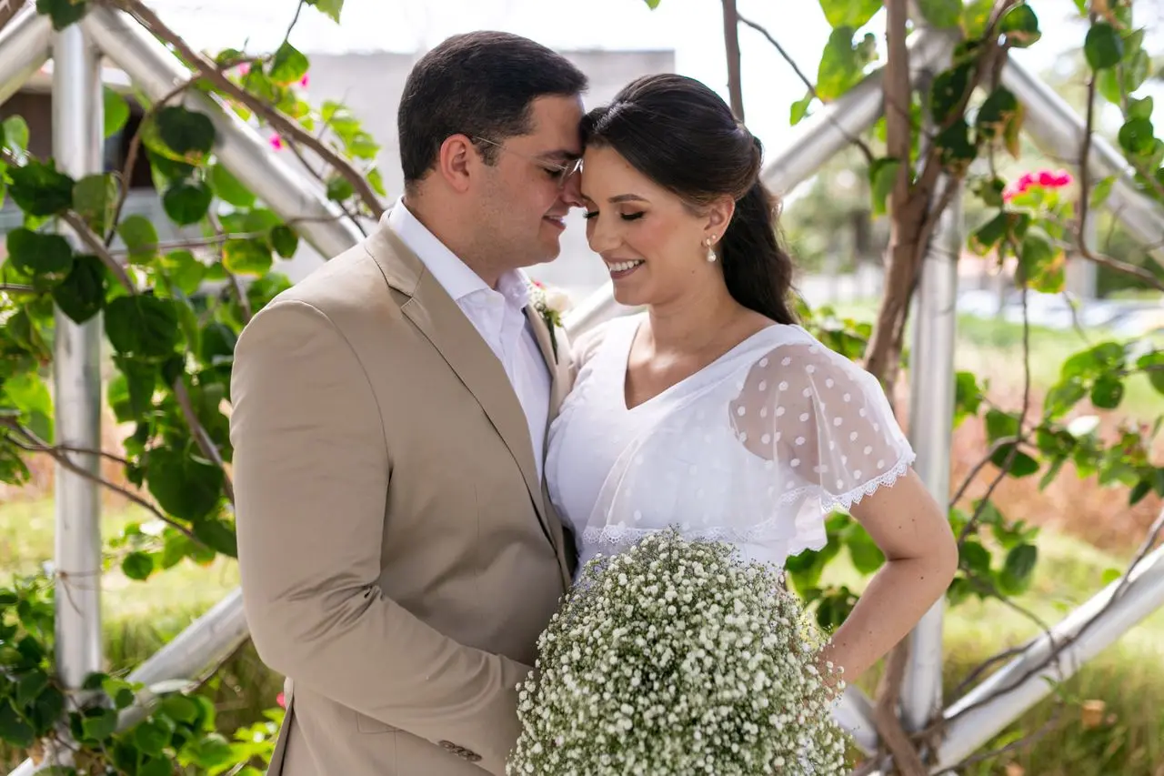 Como economizar no casamento desde os primeiros preparativos? Casal de noivos sorrindo durante cerimônia de casamento ao ar livre, com buquê de mosquitinho e decoração com folhagens.