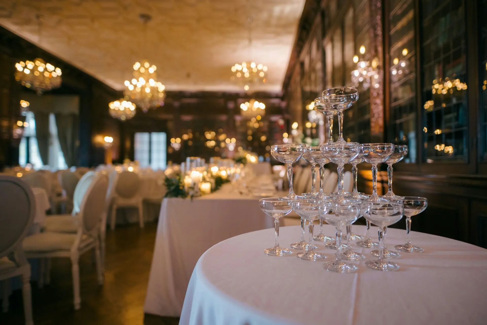 Taças de champanhe empilhadas sobre uma mesa em um elegante salão de festas decorado para casamento ou evento, com mesas, cadeiras e lustres ao fundo.
