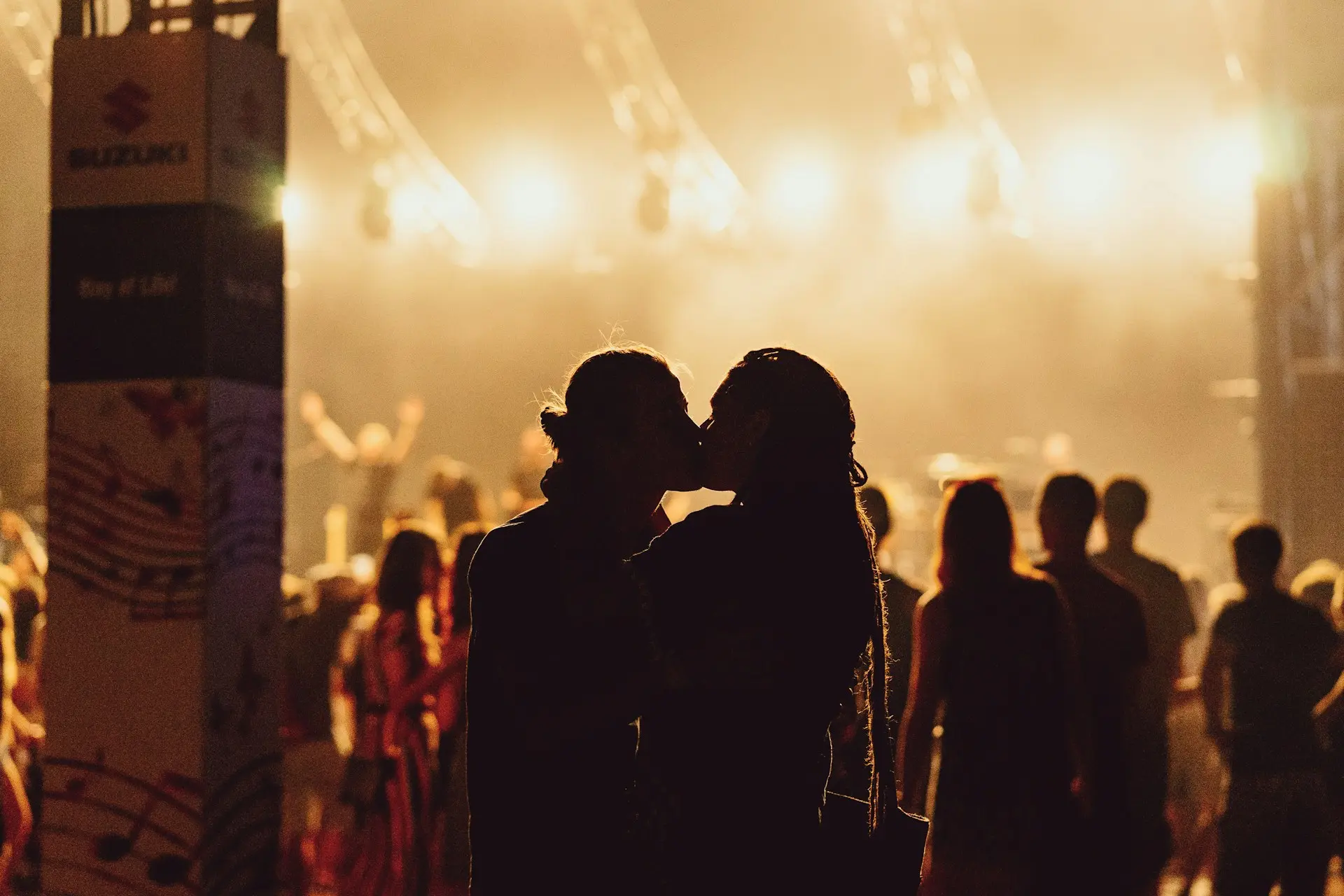 Casal se beijando durante um show, com luzes do palco ao fundo e público ao redor.