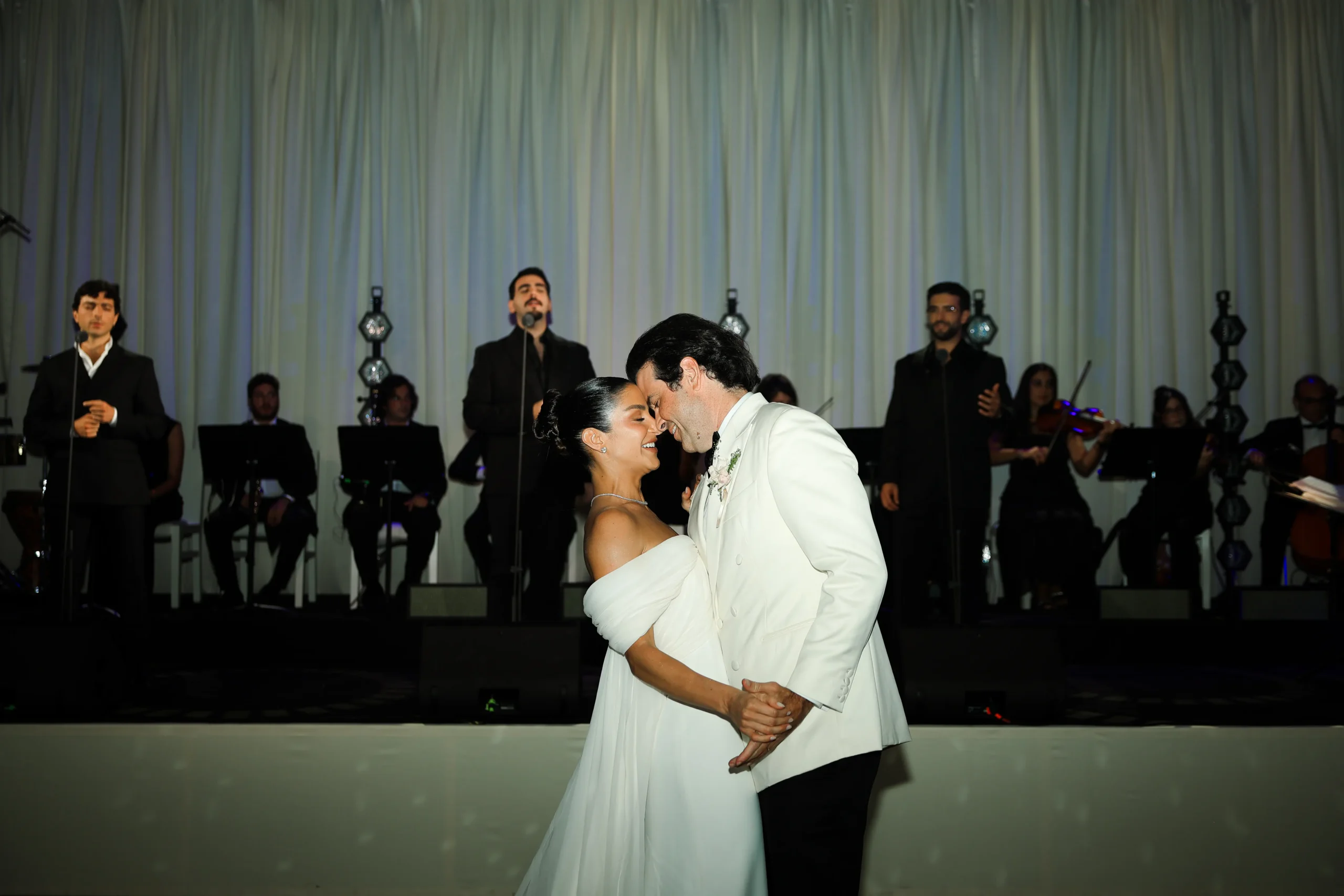 Noivos dançam juntos durante a festa de casamento, com banda e músicos ao fundo.