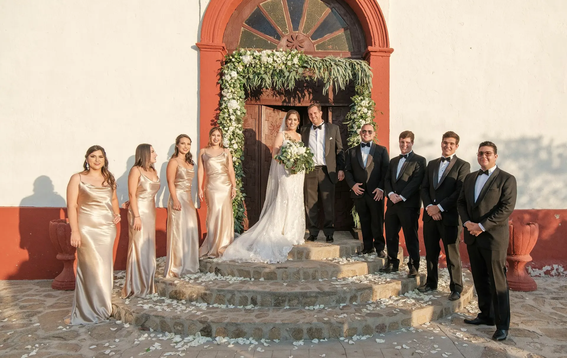 Noivos acompanhados de padrinhos e madrinhas em frente à entrada decorada de uma igreja após a cerimônia de casamento.