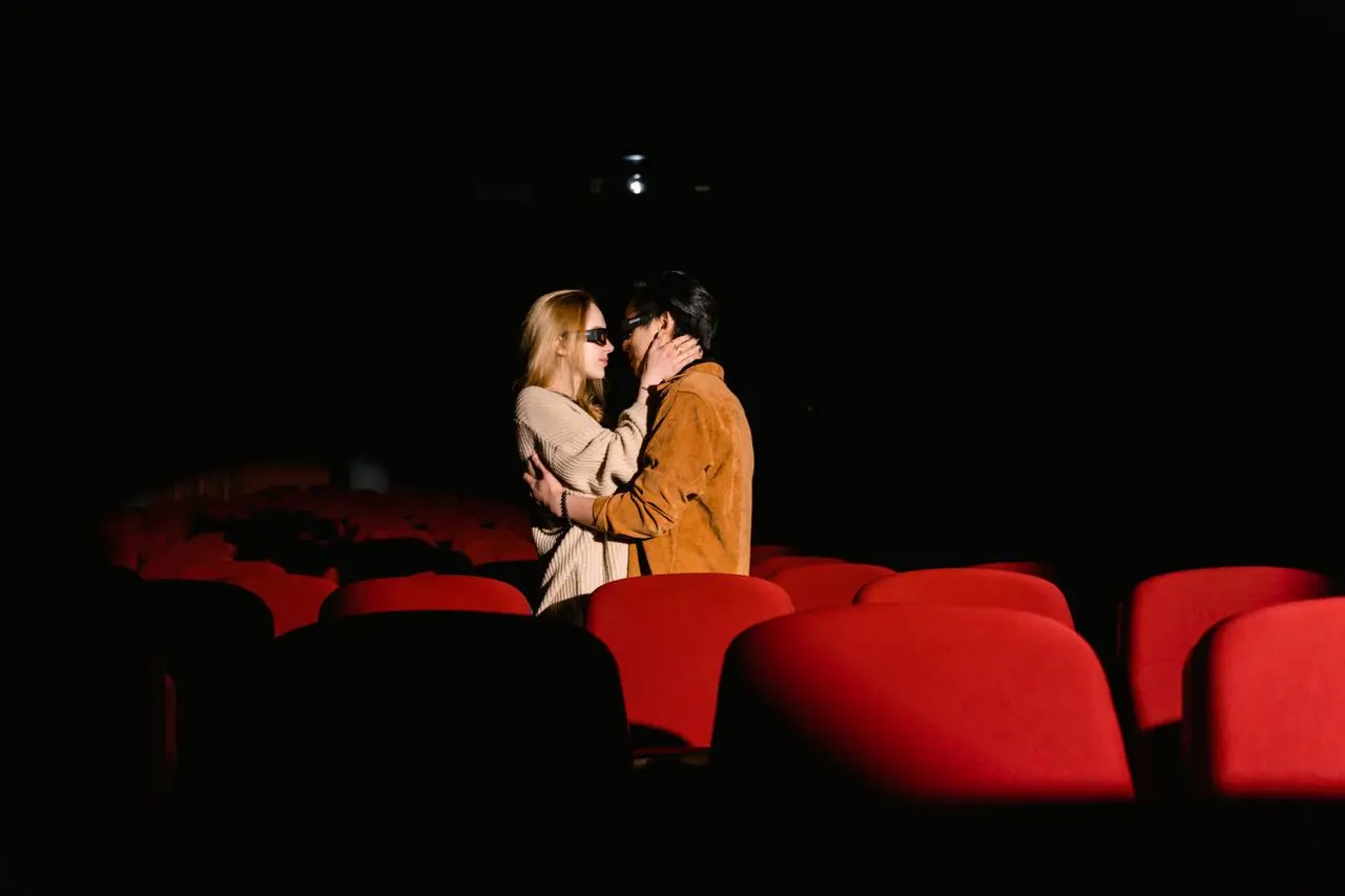 Casal em momento romântico entre as poltronas de uma sala de cinema.