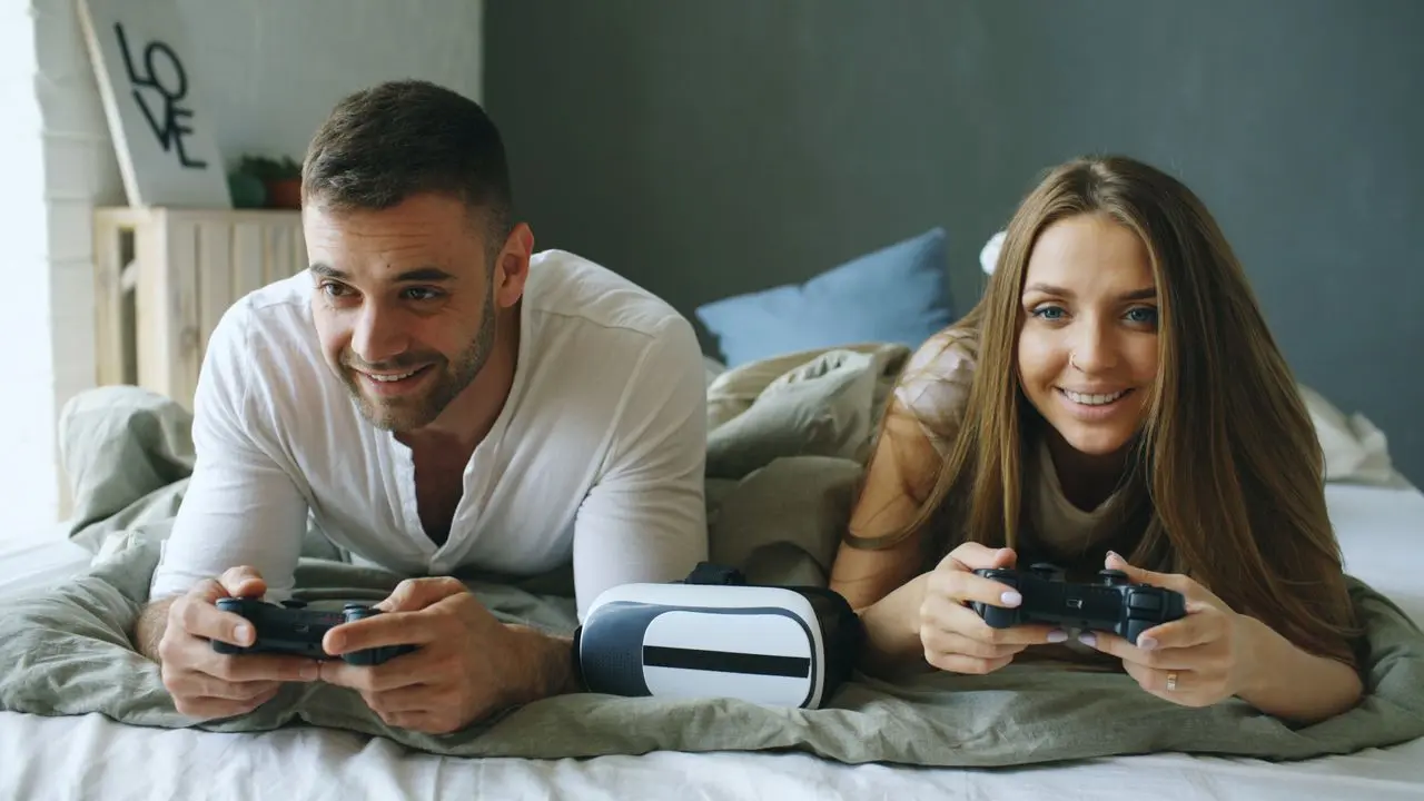 Casal jogando videogame com controles enquanto relaxa na cama.