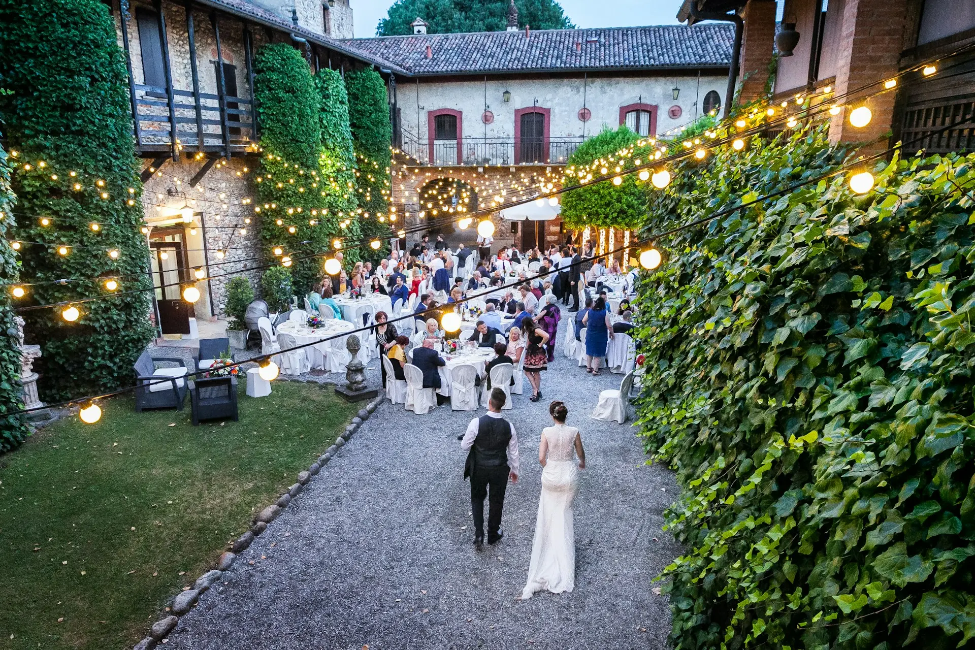 casamento-area-externa Recepção de casamento em restaurante com área externa, mesas para convidados, iluminação suspensa e pátio cercado por construções rústicas e folhagens.