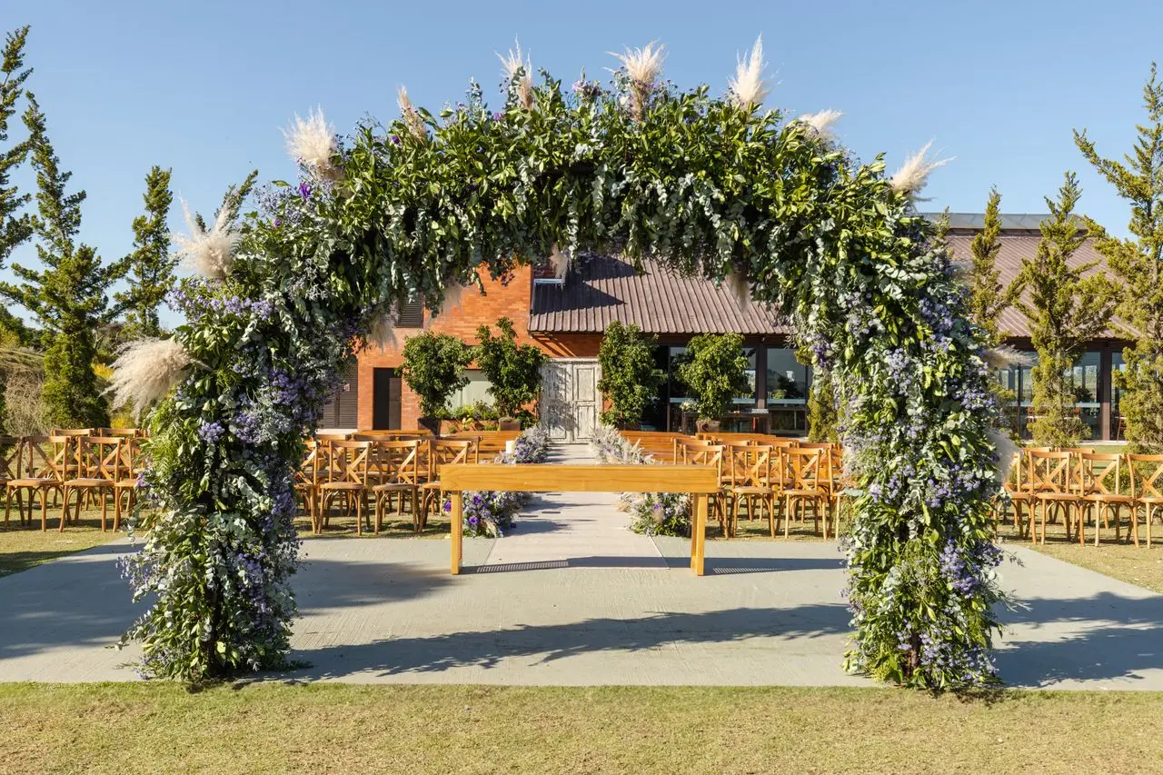 casamento-chacara Espaço para casamento em chácara ao ar livre, com altar em arco decorado com flores lilases e folhagens, cadeiras de madeira e gramado.