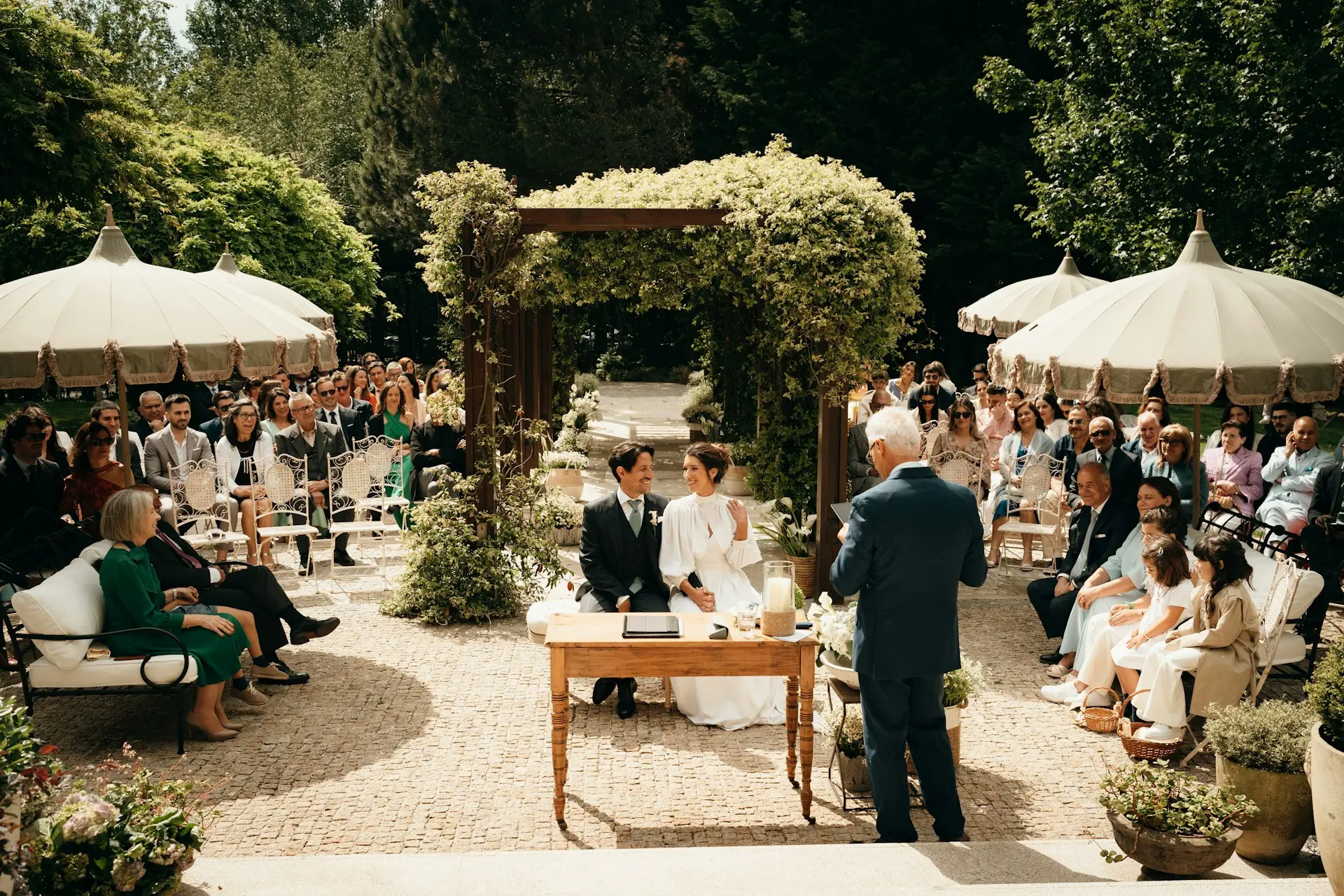 casamento-em-jardim Cerimônia de casamento em jardim ao ar livre, com noivos sentados diante de um altar decorado com flores brancas e convidados ao redor.