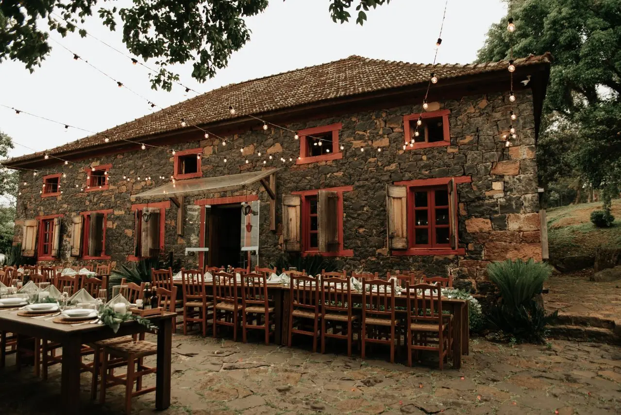 casamento-fazenda-historica Espaço para casamento em fazenda histórica, com casarão de pedra, mesas de madeira decoradas e iluminação suspensa em ambiente rústico.