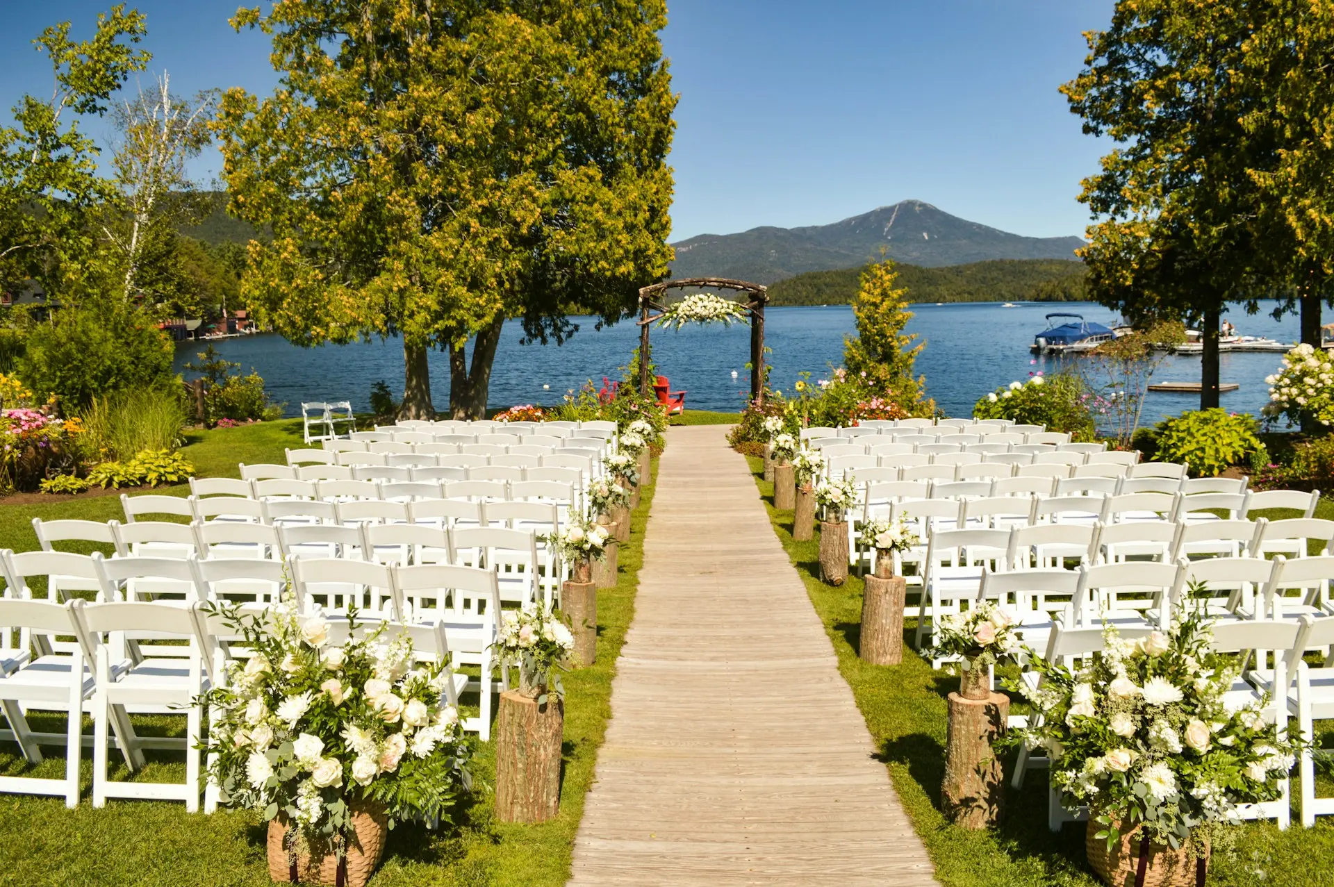 casamento-vista-para-o-mar Espaço para cerimônia de casamento ao ar livre à beira de um lago, com cadeiras brancas, corredor de madeira e altar decorado com flores.