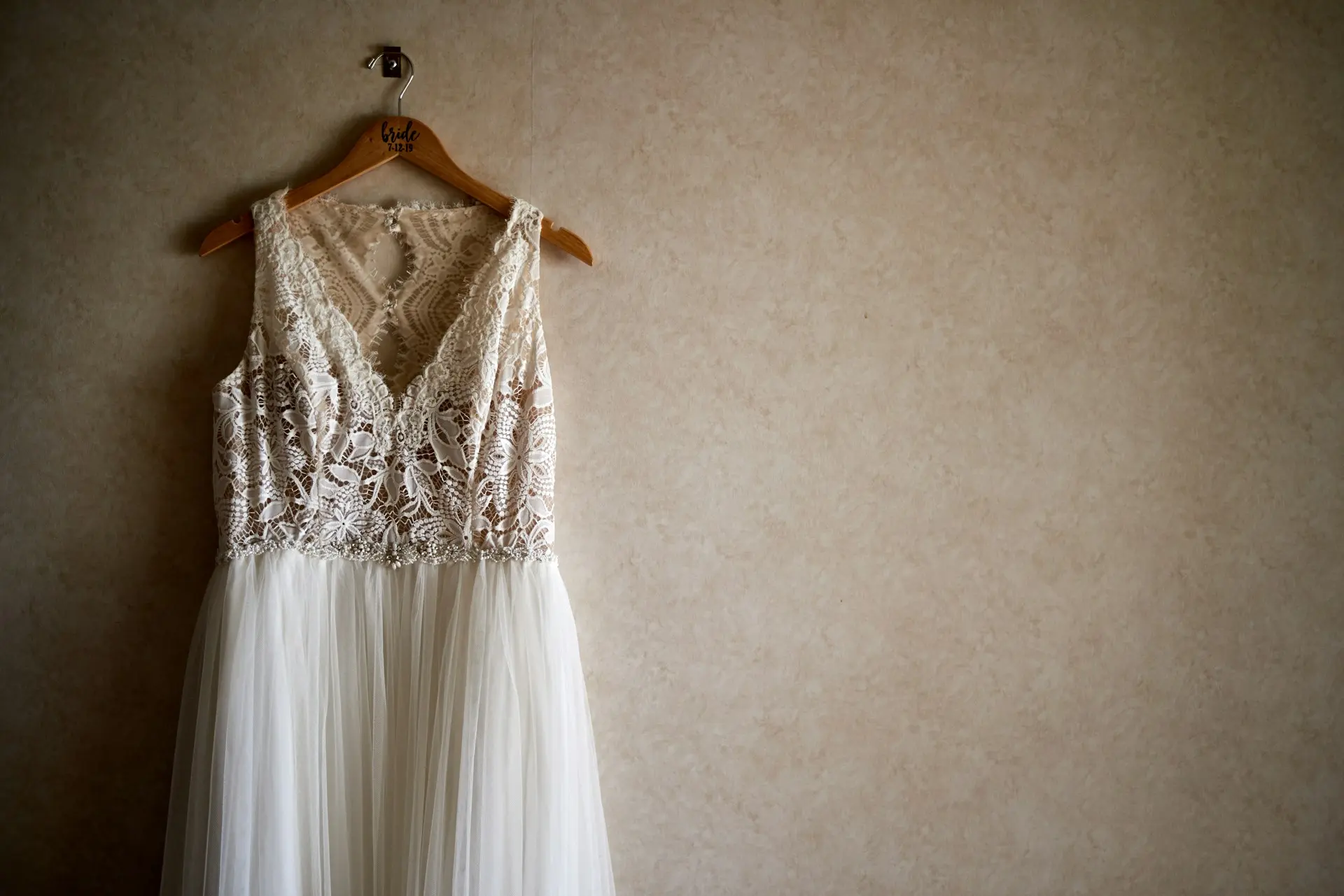 Vestido de noiva branco com corpete de renda floral, decote em V, alças largas e saia fluida de tule, pendurado em cabide de madeira.