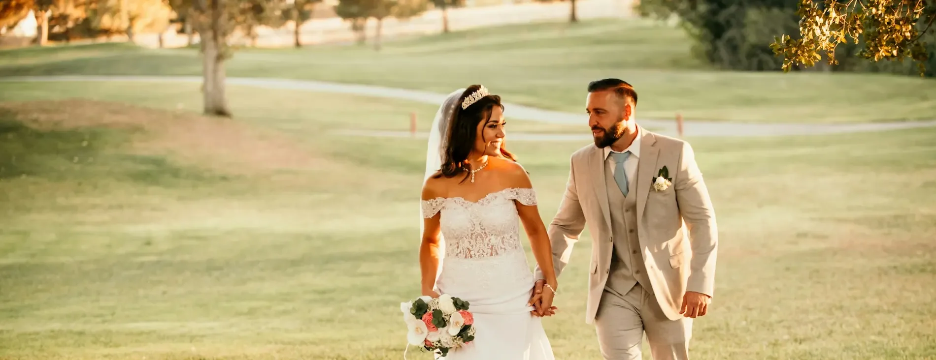 Casal de noivos caminhando de mãos dadas em um gramado ao pôr do sol, com a noiva usando vestido branco de renda e o noivo de terno bege.