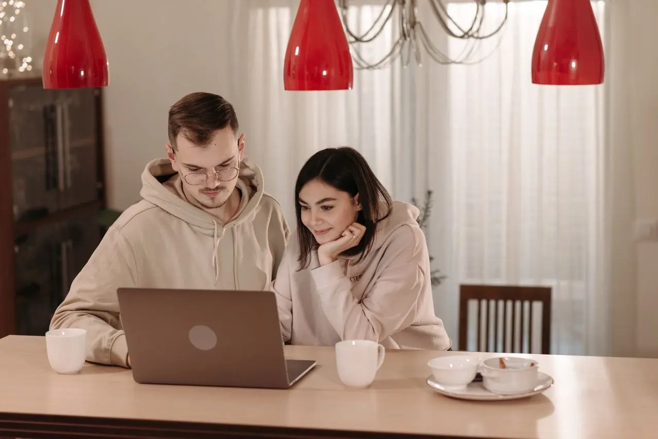 Casal sentado à mesa de casa consulta informações em um notebook, lado a lado, enquanto conversa.