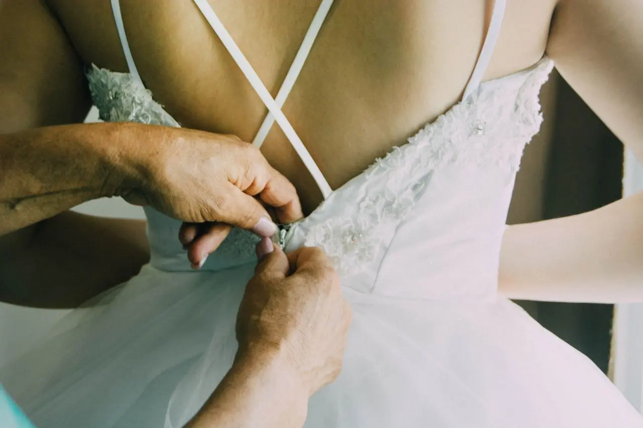Detalhe das costas de vestido de noiva branco, com alças finas cruzadas, aplicações de renda e saia de tule, sendo ajustado pelo zíper.
