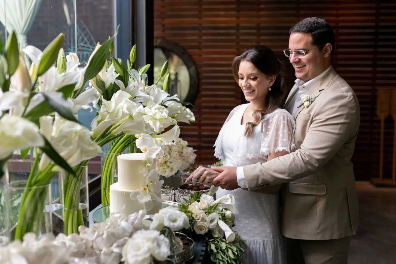 s_MG_8164 Casal de noivos sorridente corta o bolo de casamento em uma recepção intimista, com decoração de flores brancas.