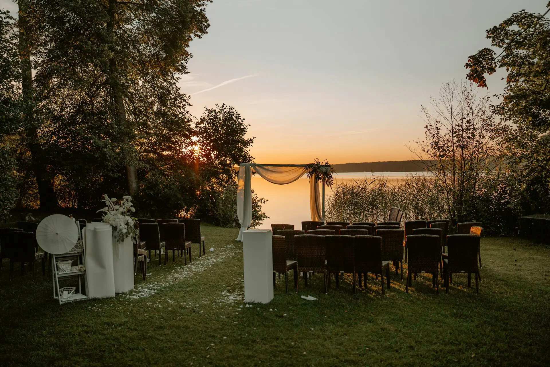 Cerimônia de casamento ao ar livre em um jardim à beira de um lago, com cadeiras para convidados, altar decorado com tecidos e flores e pôr do sol ao fundo.