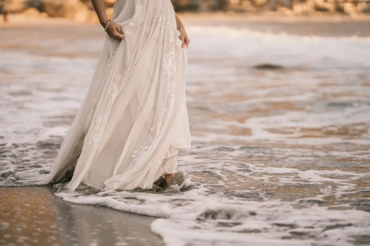 Detalhe de vestido de noiva em chiffon branco, com saia longa e fluida, tecido leve e aplicações florais, tocando a água do mar na praia.