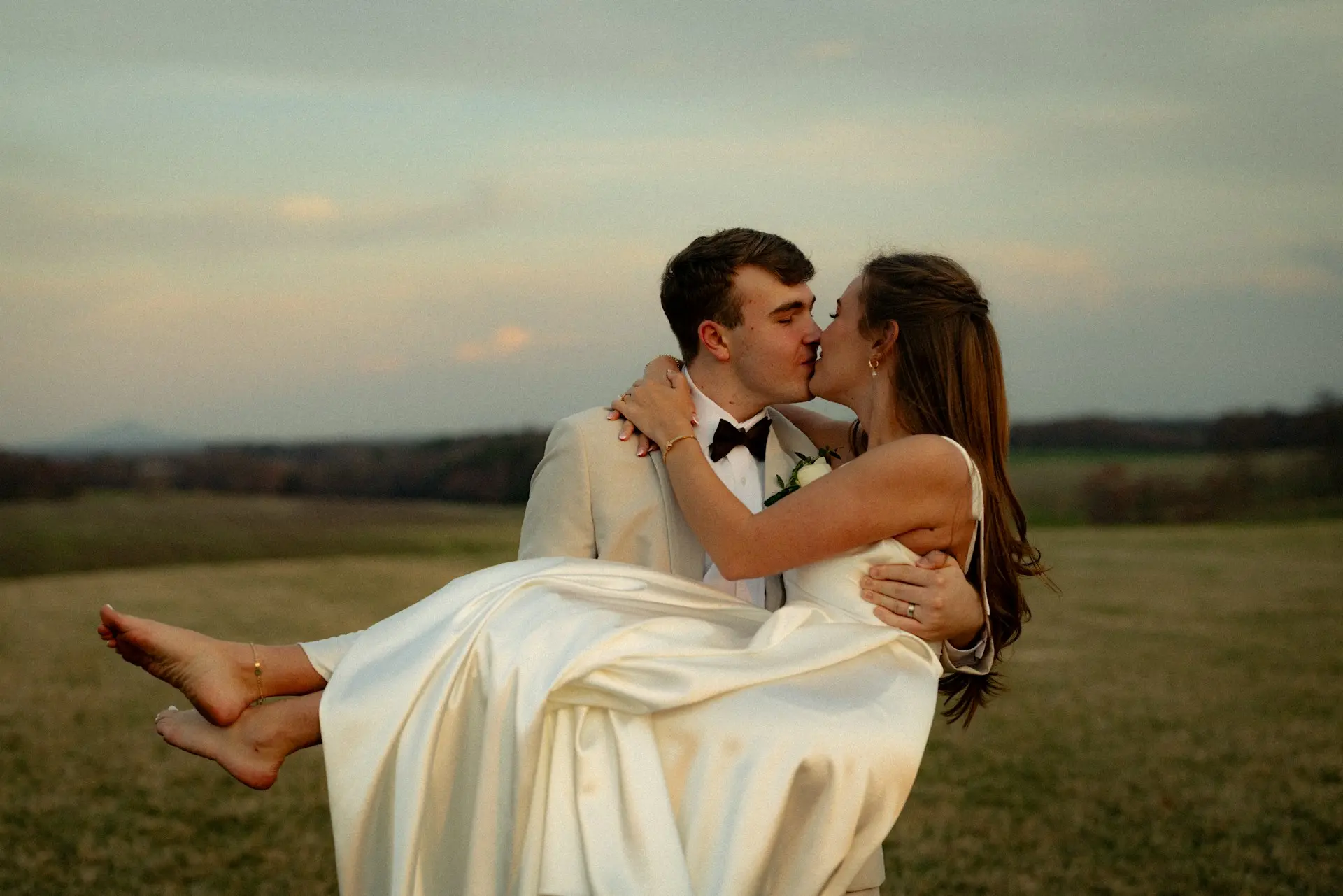 Noivo de terno bege segurando a noiva no colo enquanto se beijam em um campo ao pôr do sol. A noiva usa vestido de casamento de cetim branco, com saia longa e fluida.
