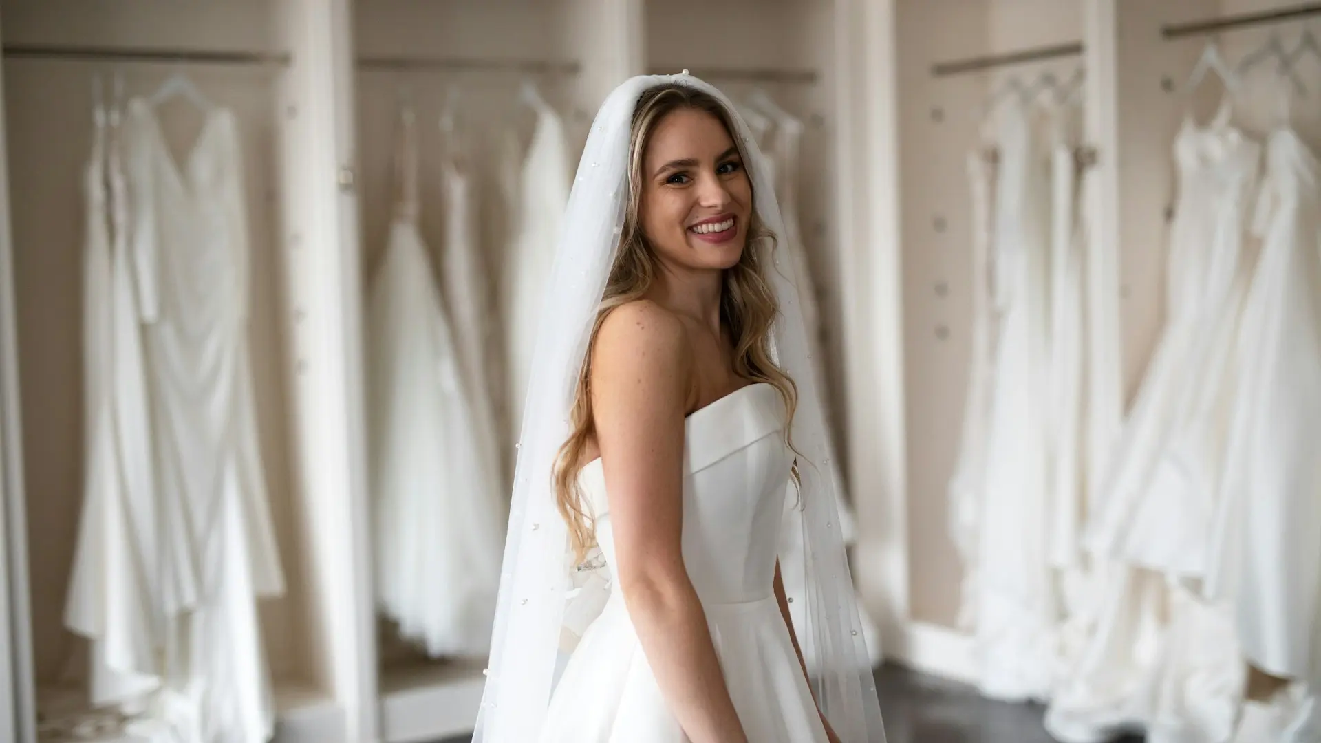 Noiva usando vestido de casamento em crepe branco, com corpete estruturado, decote tomara que caia e saia lisa, acompanhada de véu longo.