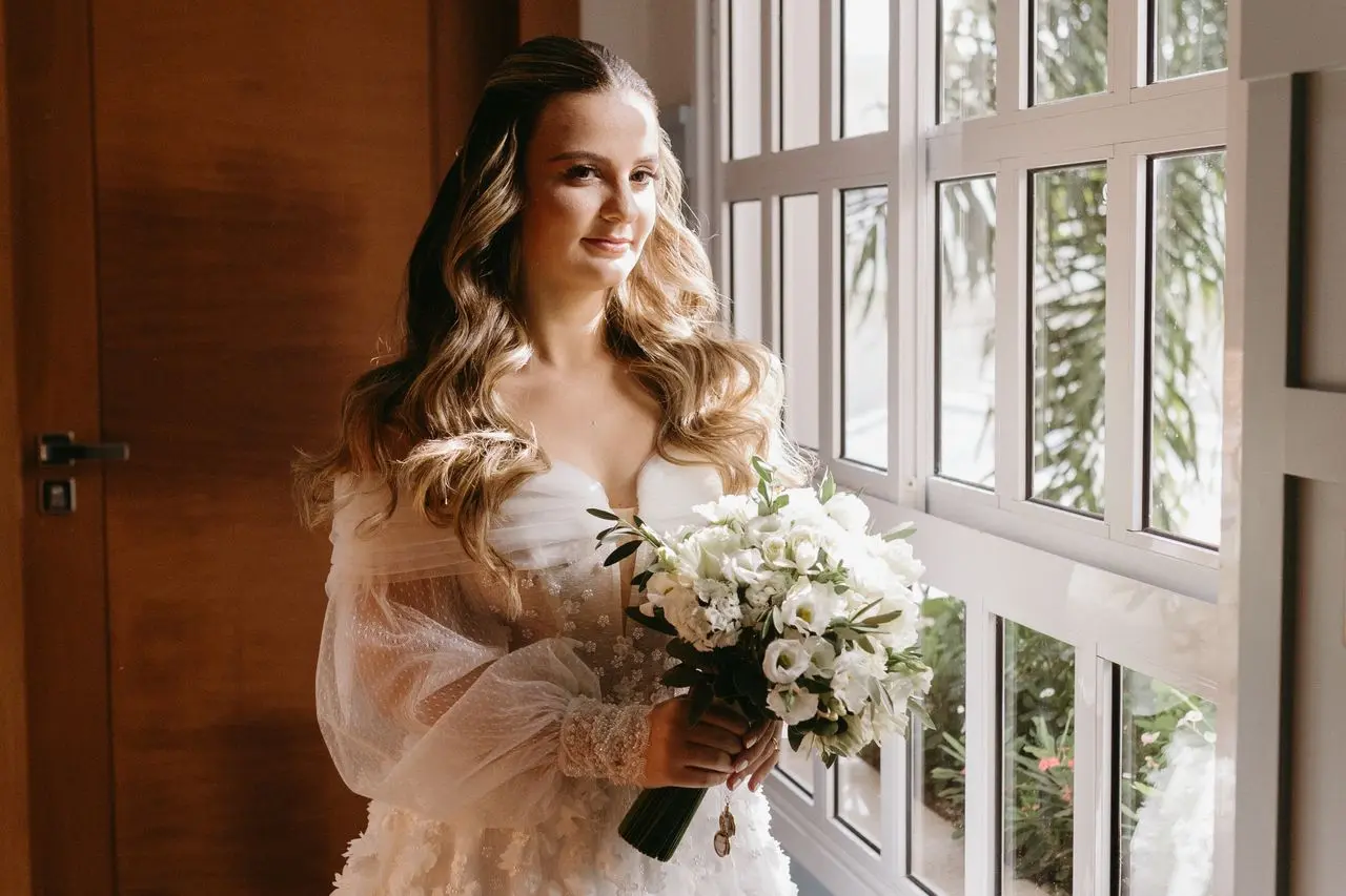 Noiva com vestido de casamento em organza branca, com mangas bufantes transparentes, detalhes florais e decote ombro a ombro, segurando um buquê de flores brancas.