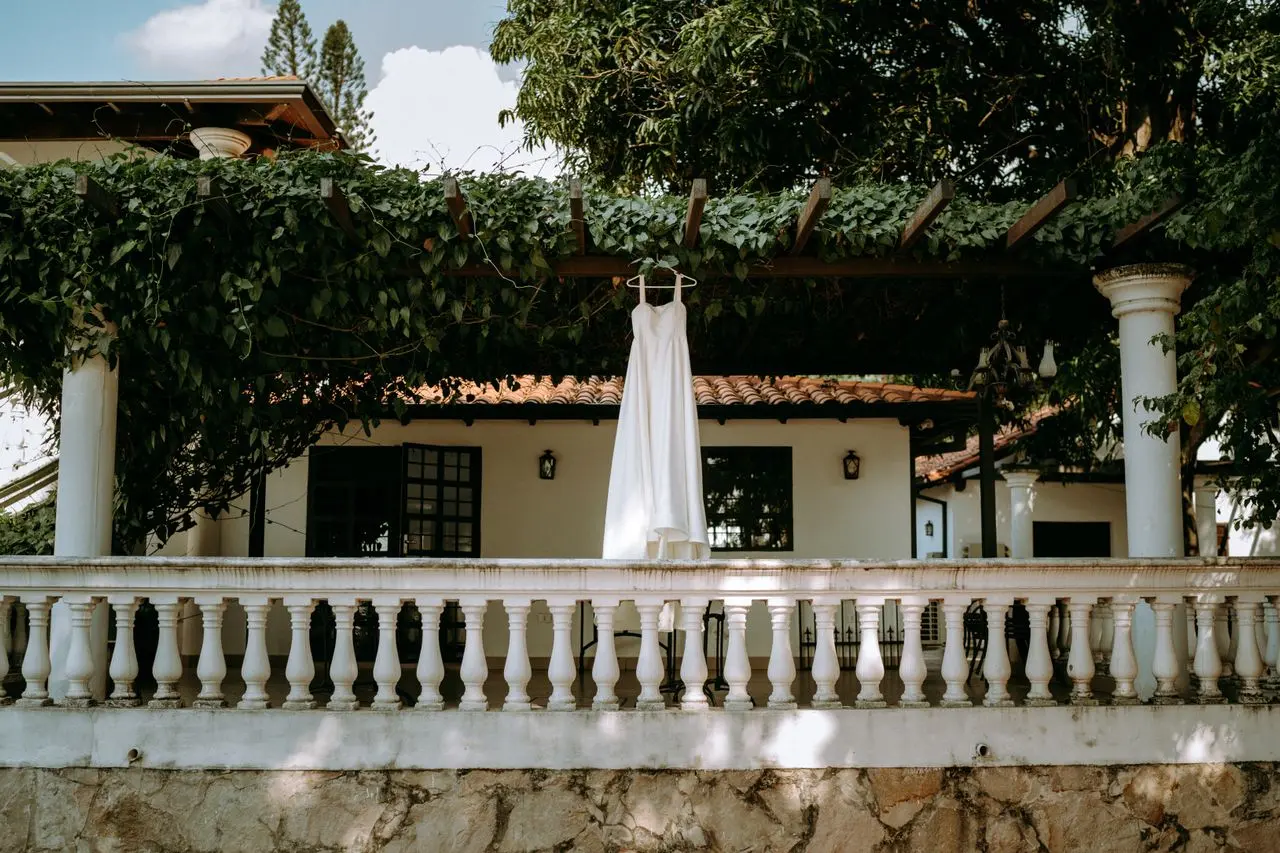 Vestido de noiva branco, longo e fluido, pendurado em um pergolado coberto por folhagens, em área externa de uma casa rústica.