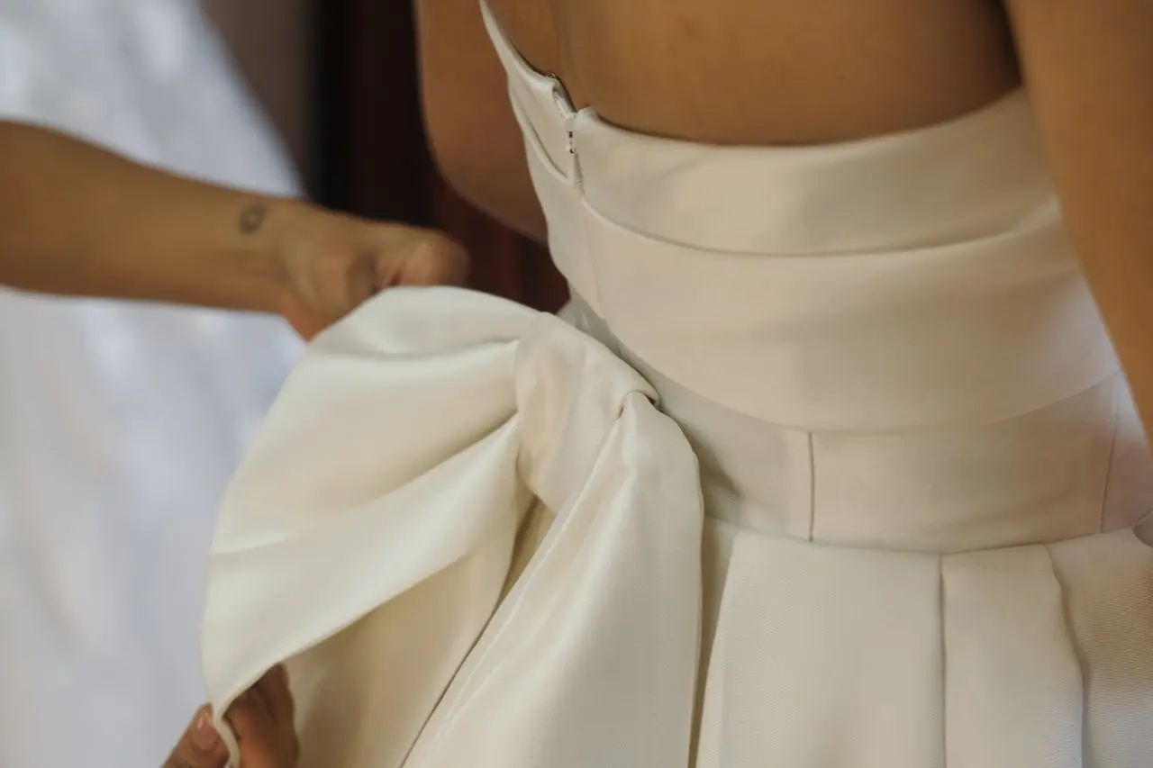 Detalhe de vestido de noiva em tecido zibeline branco, com estrutura encorpada, cintura marcada e pregas volumosas sendo ajustadas.