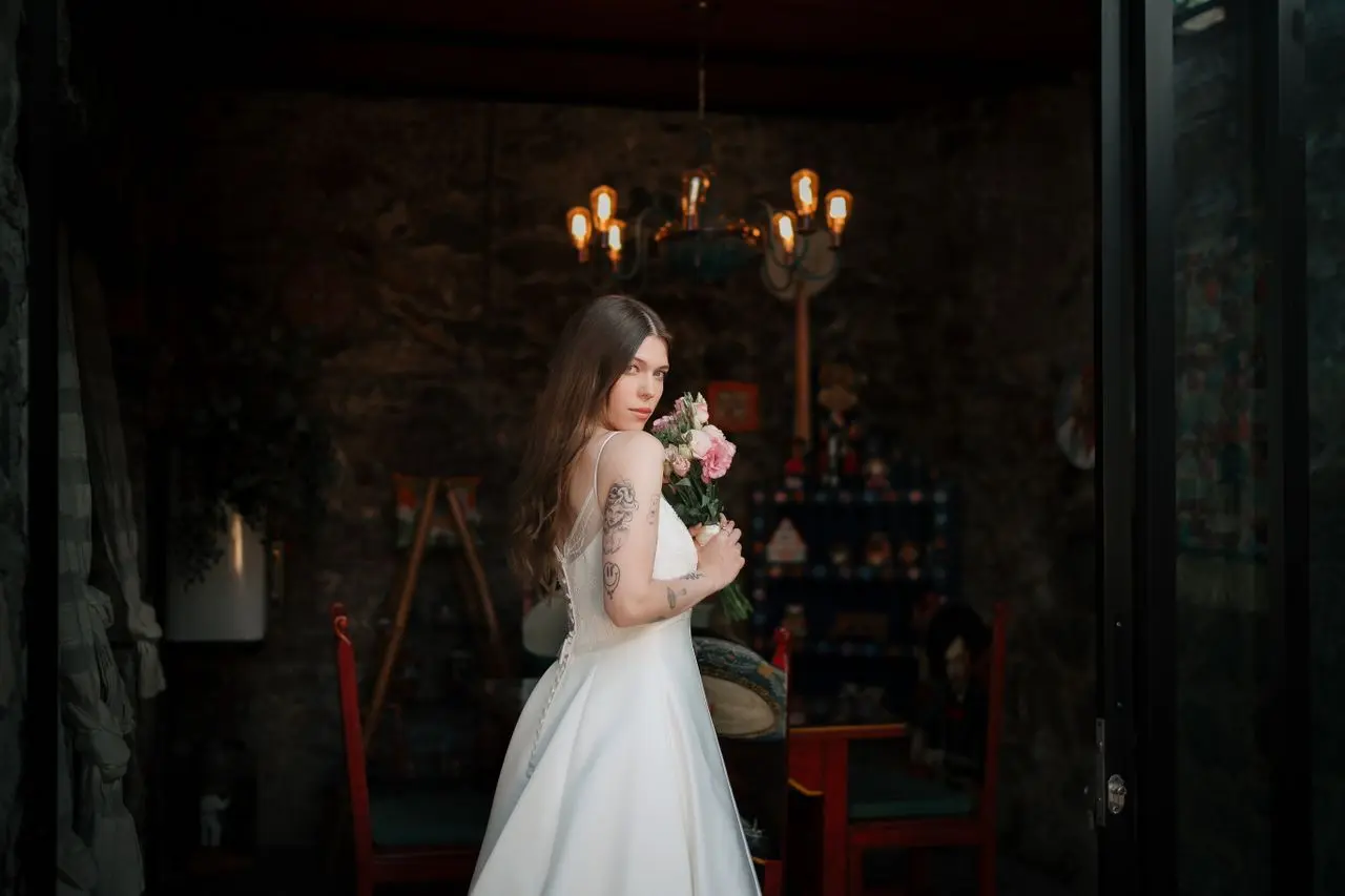 Noiva com vestido de casamento evasê branco, saia ampla e alças finas, segurando um buquê de flores em ambiente rústico.