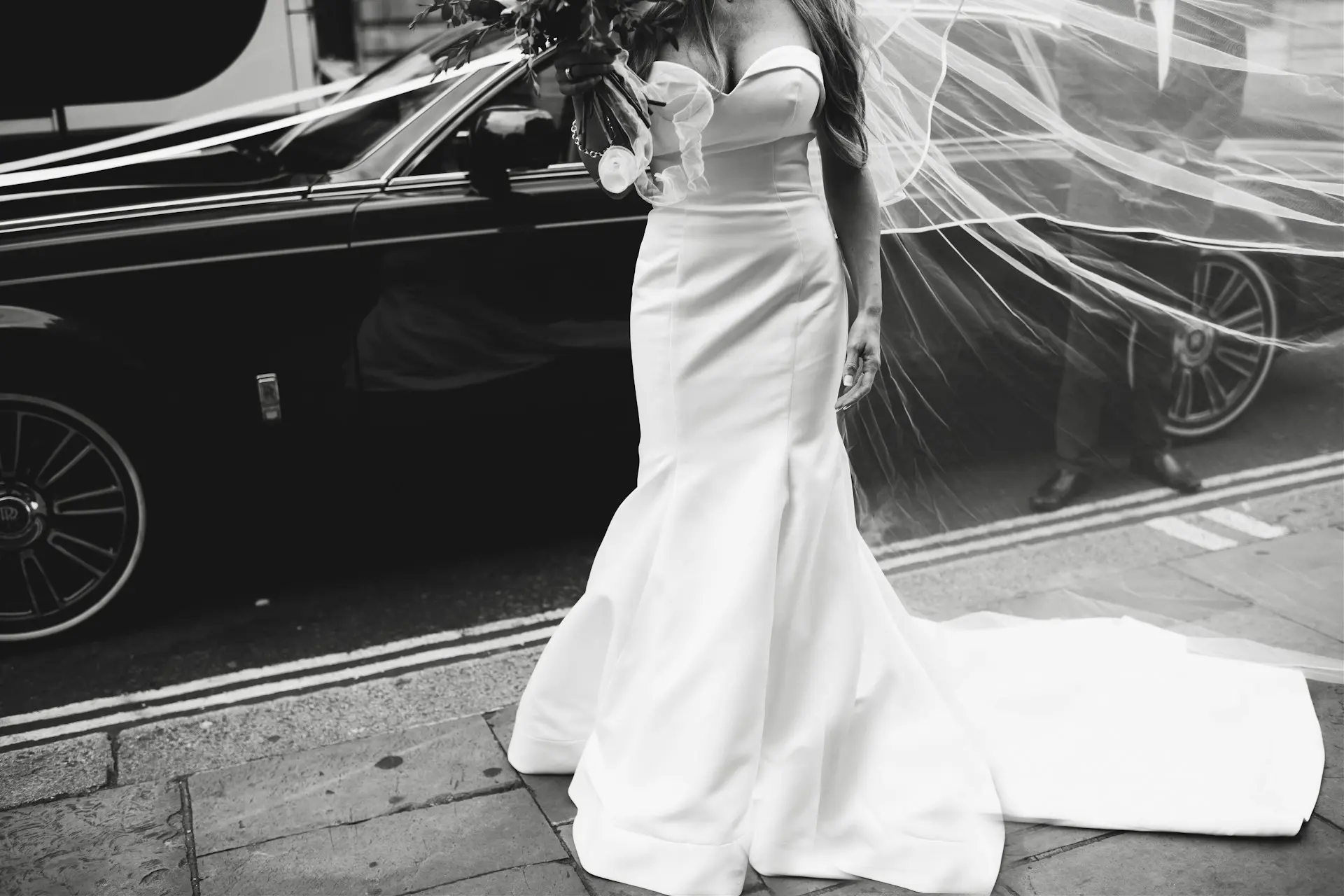 Noiva com vestido de casamento sereia branco, ajustado ao corpo, com ombros à mostra, saia ampla a partir dos joelhos e cauda longa.